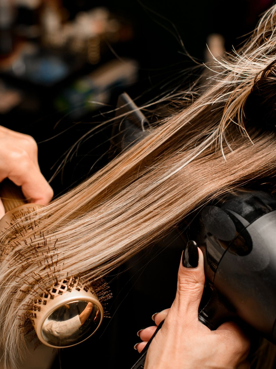 Une coiffeuse sèche des cheveux blonds au sèche-cheveux avec une brosse ronde et un sèche-cheveux noir dans un salon.