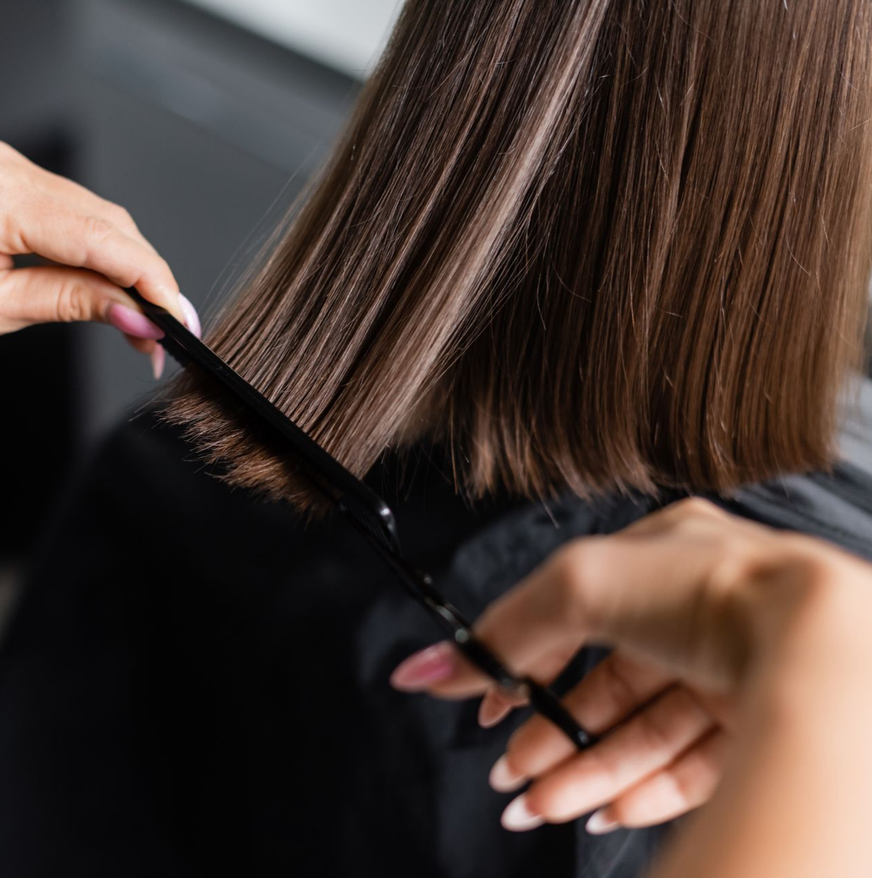 Une coiffeuse coupe les cheveux bruns et raides d'une personne avec des ciseaux et un peigne dans un salon de coiffure.