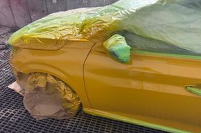Ein gelbes Auto, teilweise mit Plastikfolie und Klebeband abgeklebt, befindet sich in einer Lackierkabine.