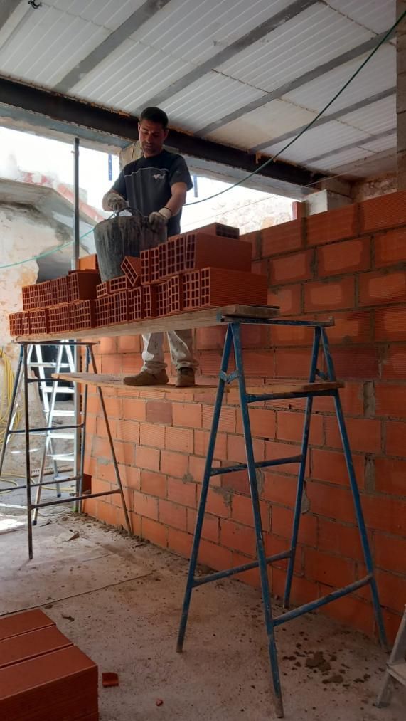Un hombre coloca ladrillos en un andamio dentro de un edificio. Lleva guantes y trabaja en una pared de ladrillos rojos.