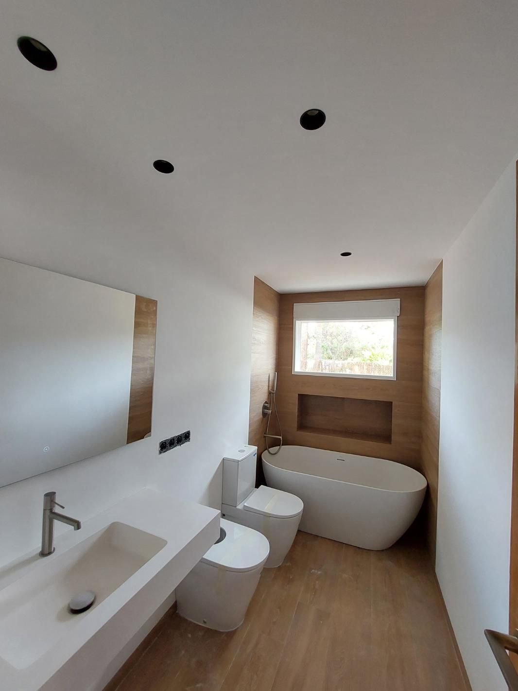 Baño moderno con accesorios blancos, bañera independiente y pared con acento en tono madera.