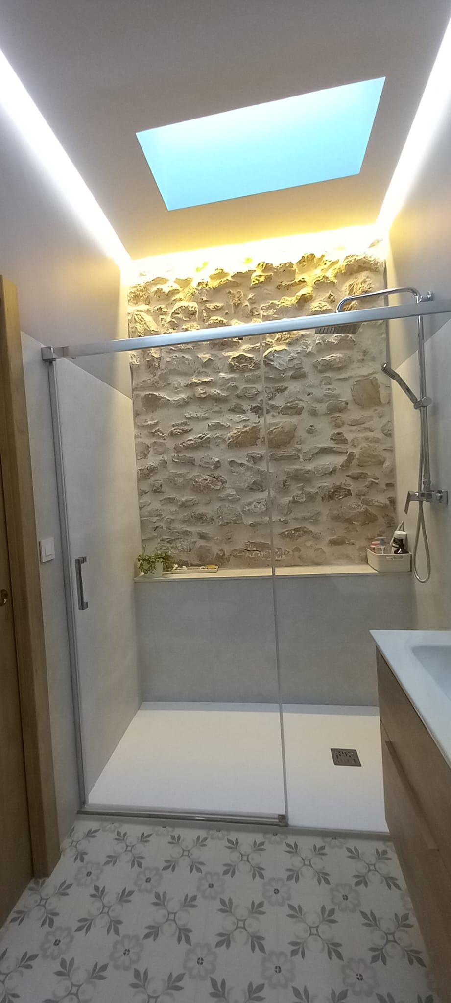 Baño con pared de piedra, ducha y claraboya. Suelo de baldosas con estampado floral.