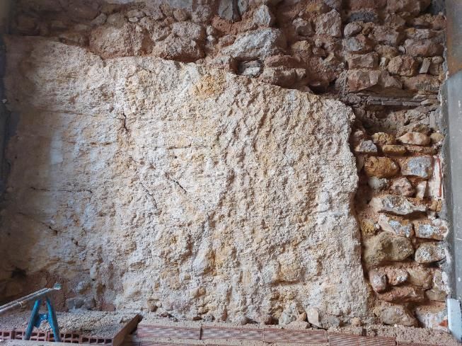 Muro de piedra vista con yeso rugoso de color marrón claro. Se aprecia un trozo de mampostería a la derecha.