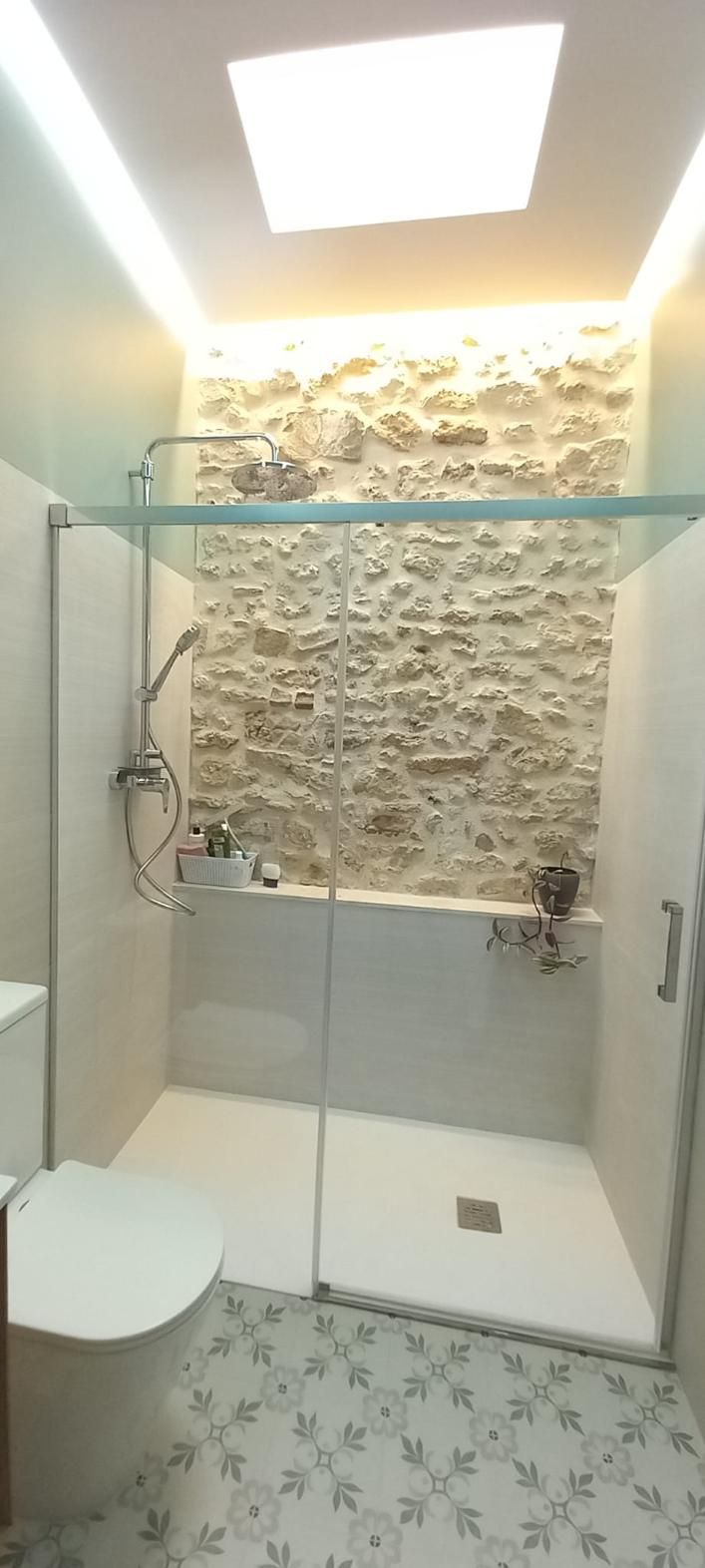 Baño con pared con acento de piedra en la ducha, puerta de vidrio, inodoro blanco y pisos de baldosas estampadas.