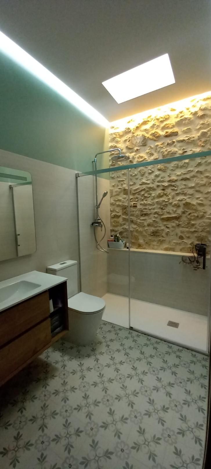 Baño con pared de piedra, ducha, inodoro y lavabo de madera. 