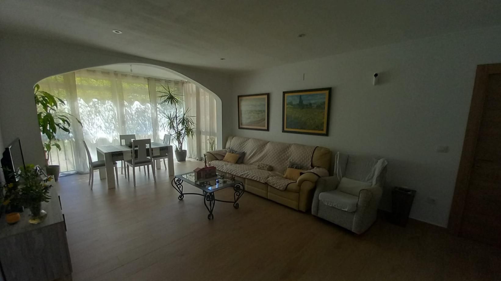 Amplia sala de estar con comedor. Paredes blancas, muebles claros y un gran ventanal arqueado.