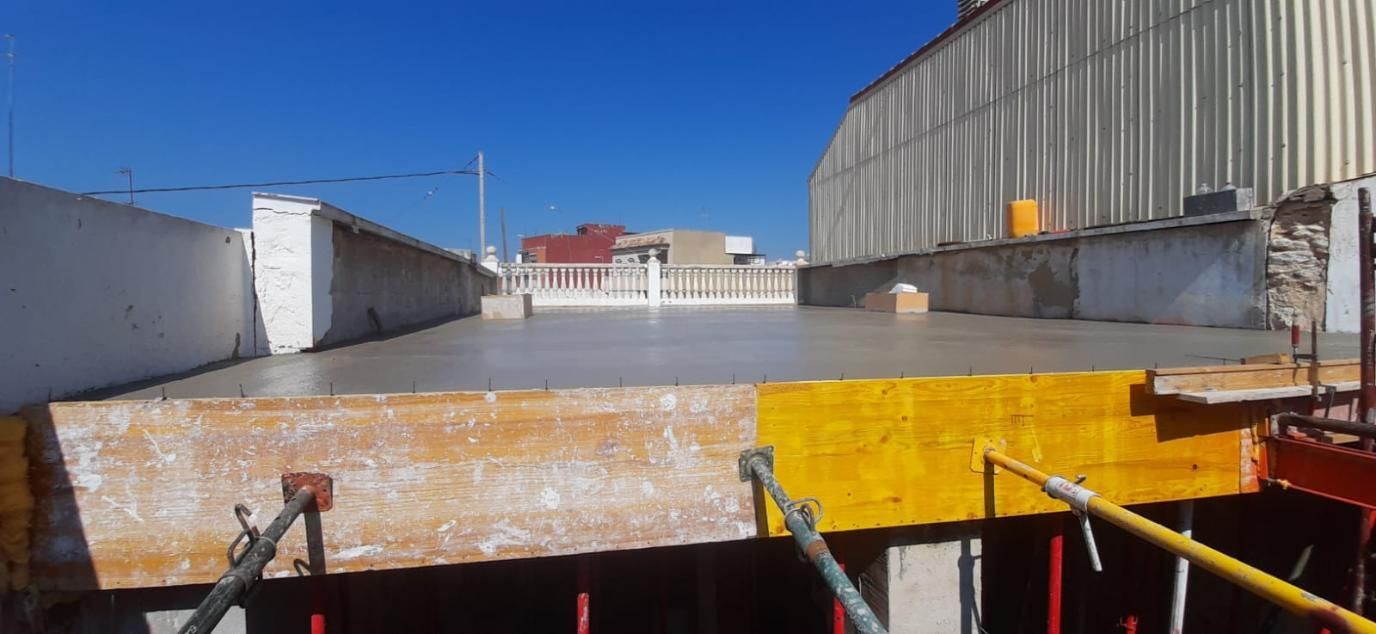 Losa de hormigón recién vertida en obra. Vigas de madera amarillas forman la estructura. Cielo azul.