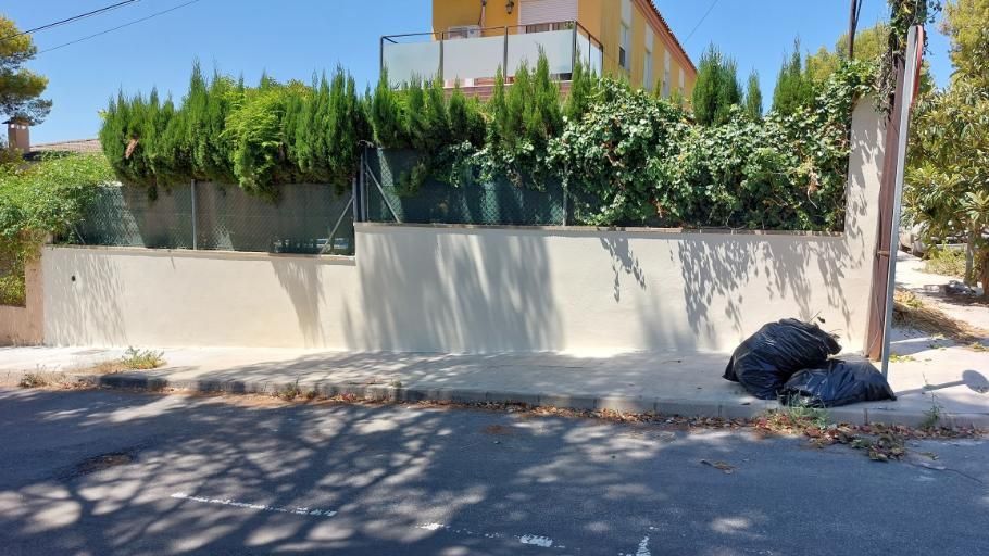 Un muro de hormigón con vegetación y bolsas de basura negras en la acera. Se ve un edificio al fondo.