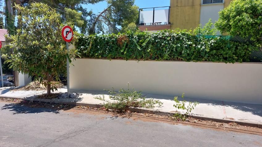 Vista de la calle: pared blanca con vegetación, sin señal de giro a la izquierda y edificio al fondo.