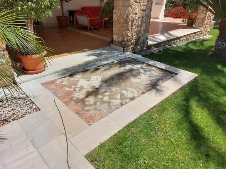 Un patio cuadrado con baldosas faltantes, rodeado de césped y un edificio con columnas.