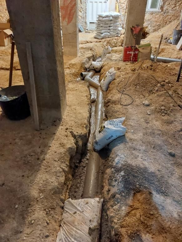 Zanja en un sitio de construcción con tuberías colocadas, sacos de arena y herramientas visibles.