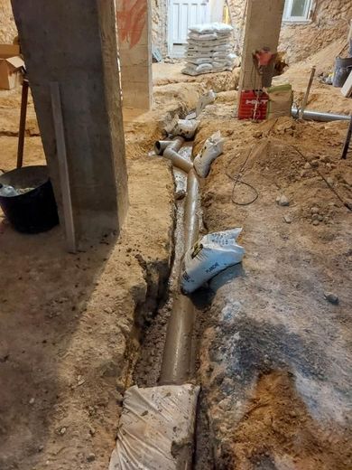 Zanja en un sitio de construcción con tuberías colocadas, sacos de arena y herramientas visibles.