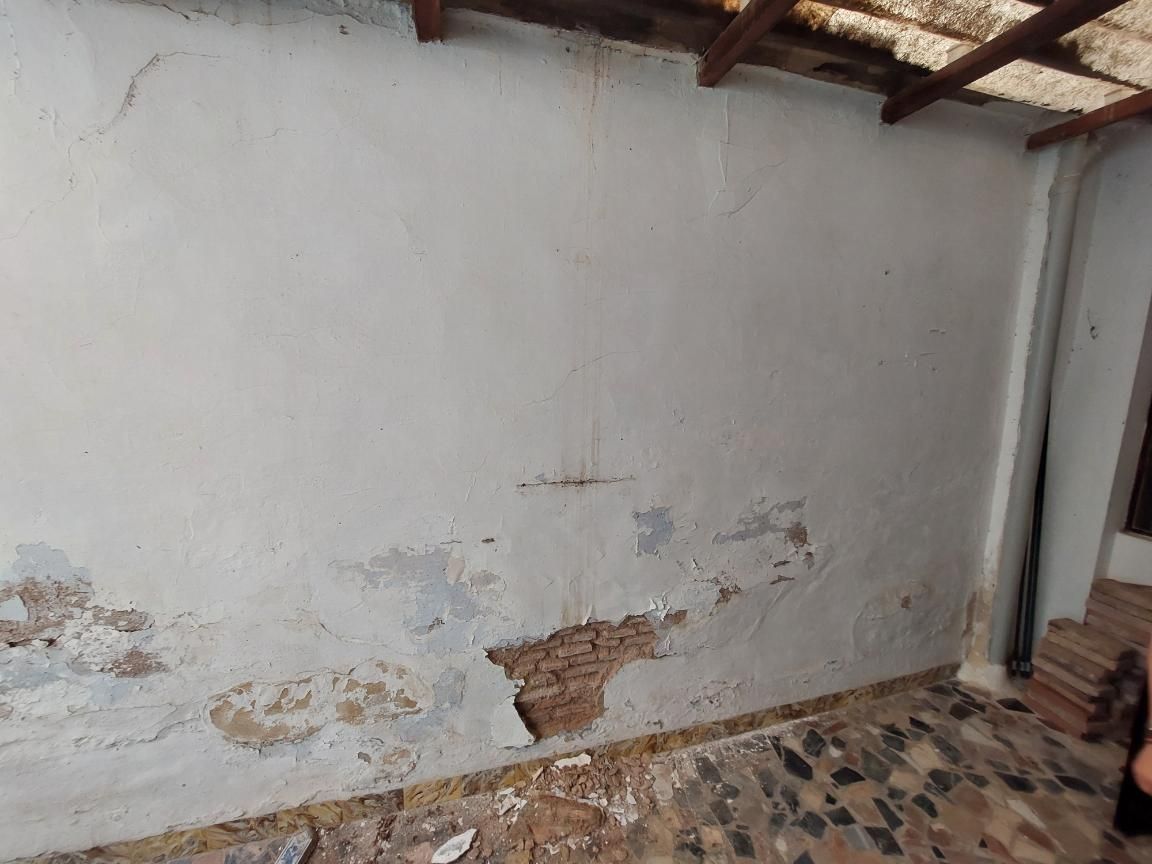 Pared blanca deteriorada con pintura descascarada, dejando al descubierto el ladrillo. Techo de madera y suelo de piedra.