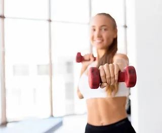 Una mujer sostiene dos mancuernas rojas sobre sus hombros en un gimnasio.