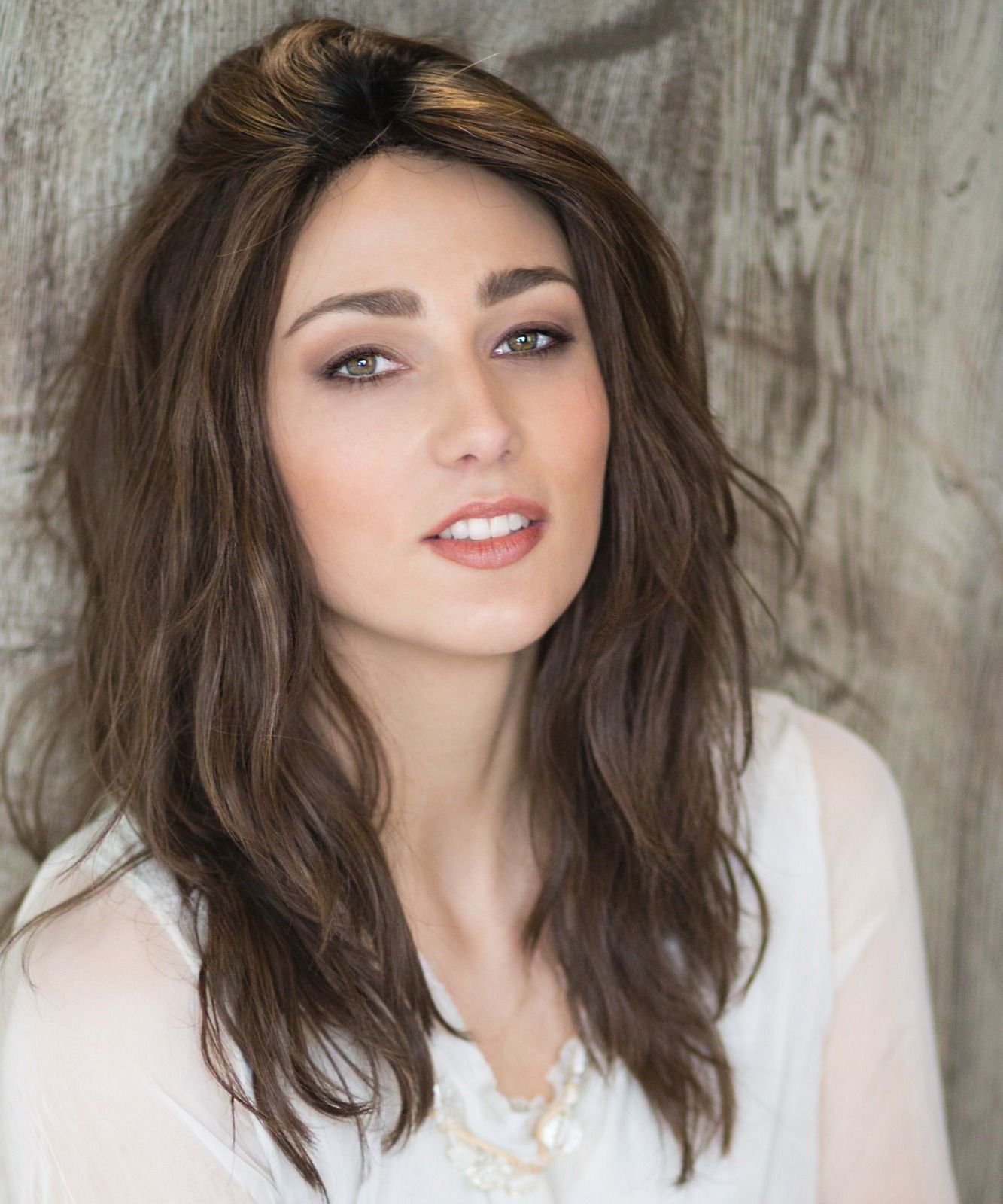 Eine Frau mit Perücke und weißem Hemd steht vor einer Holzwand.