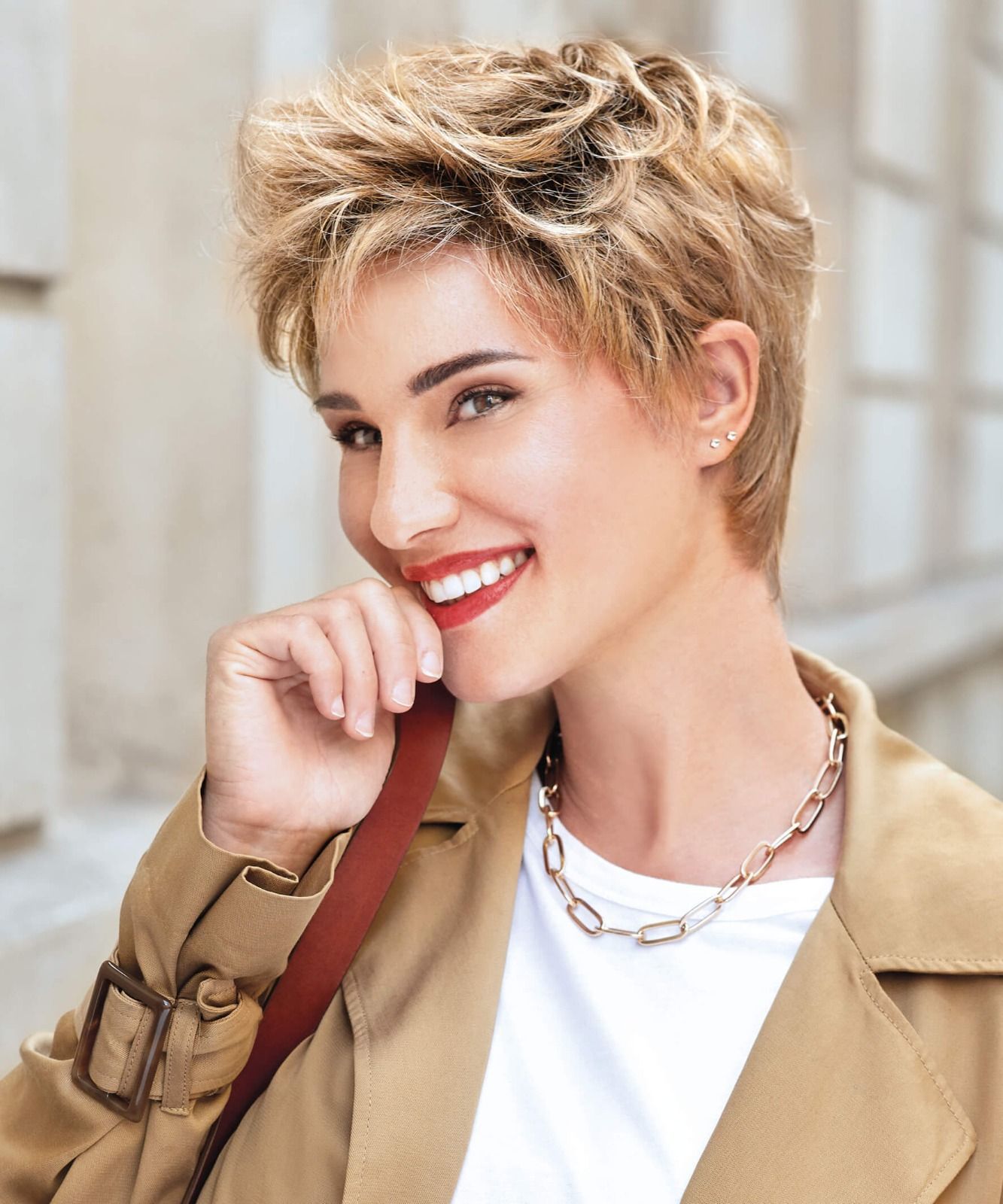 Eine Frau mit kurzen blonden Haaren trägt einen Trenchcoat und eine Halskette.