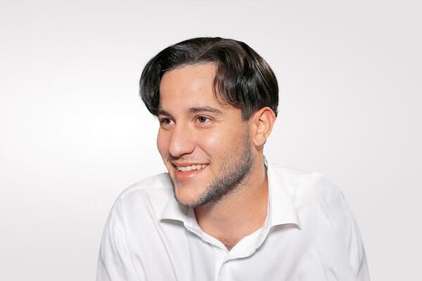 Ein junger Mann mit Bart trägt ein weißes Hemd und lächelt.