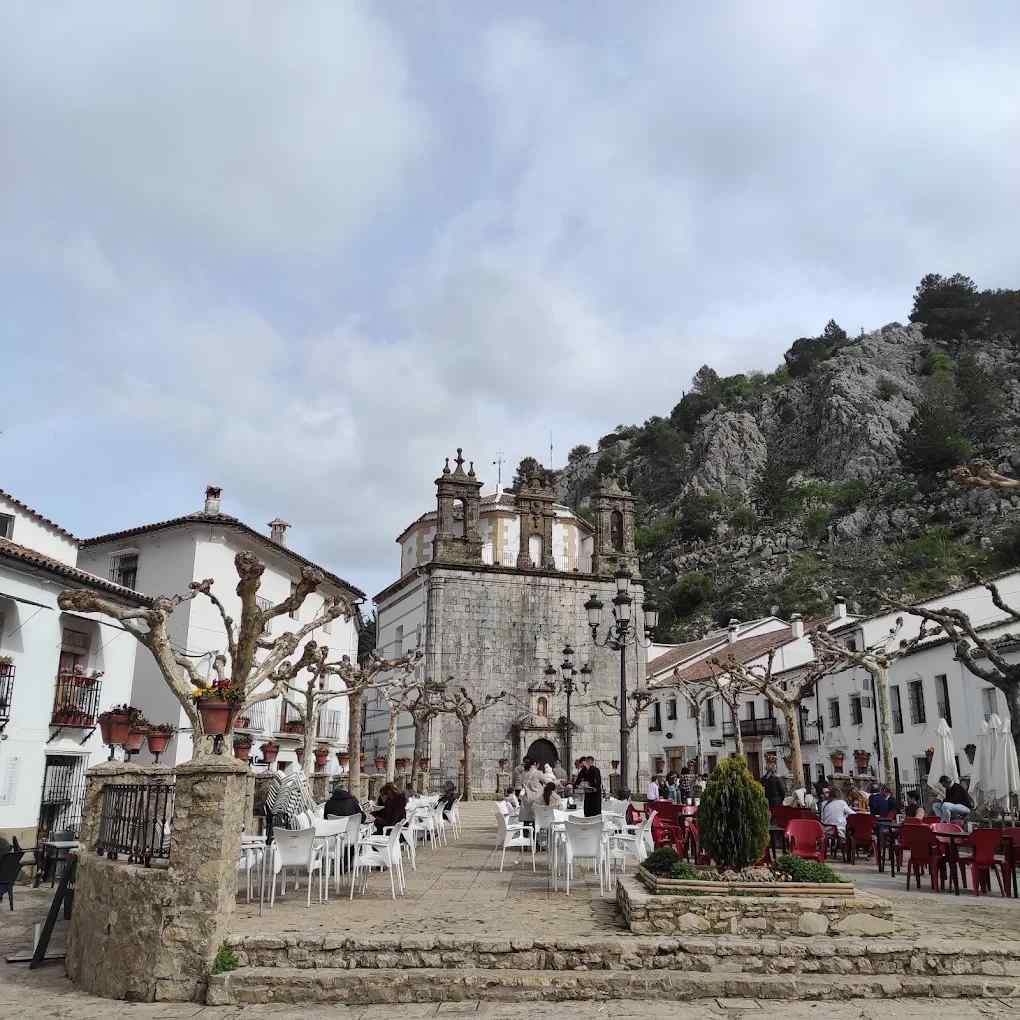 Plaza del pueblo con iglesia, mesas y sillas, gente y montañas de fondo. Cielo nublado.