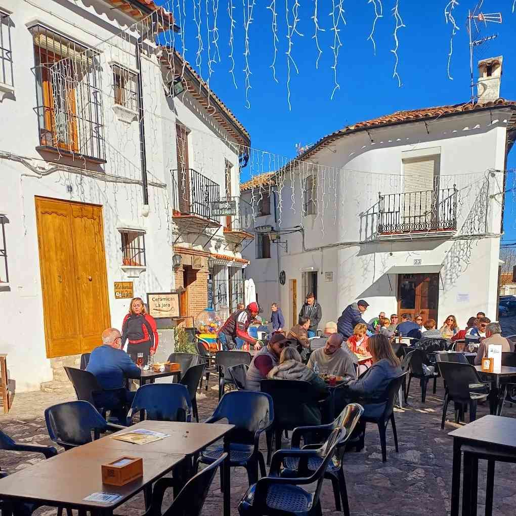 Gente en mesas de café al aire libre en una plaza de paredes blancas en un día soleado.