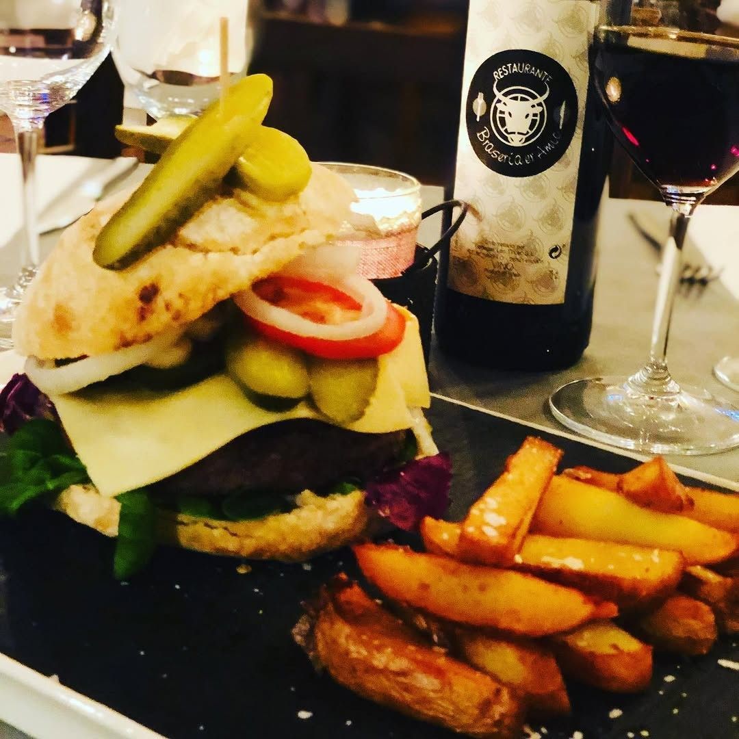 Hamburguesa alta con pepinillos y patatas fritas en una mesa de restaurante con copas de vino.