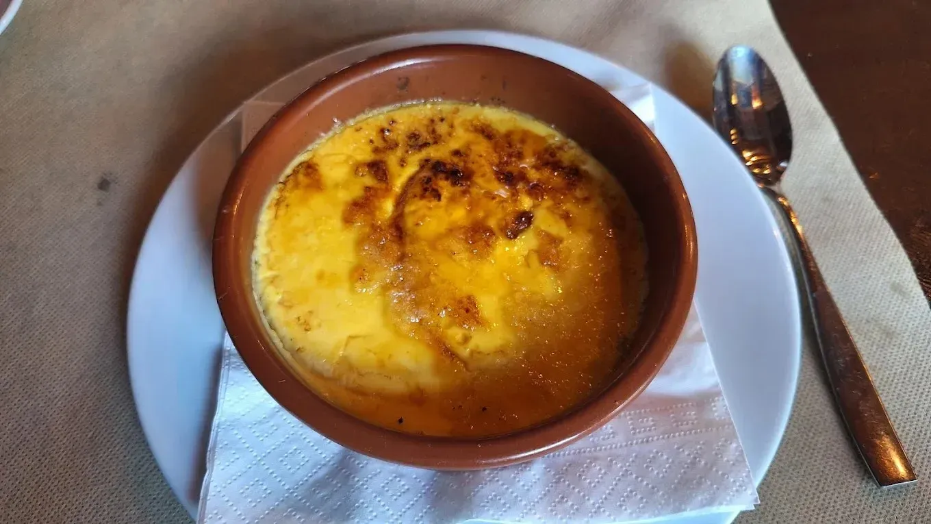 Postre de natillas horneadas en un ramequín marrón sobre un plato blanco con una cuchara sobre una servilleta.