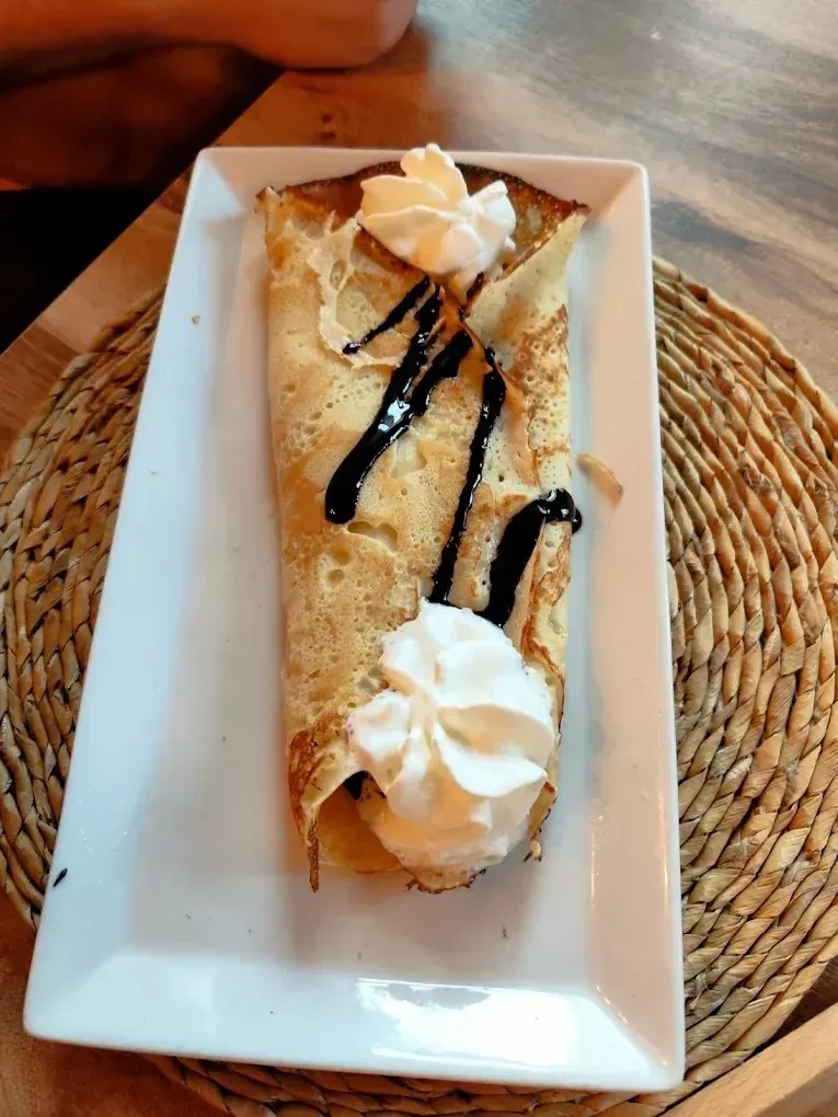 Crepe en un plato rectangular blanco, cubierto con crema batida y sirope de chocolate, sobre un mantel individual tejido.