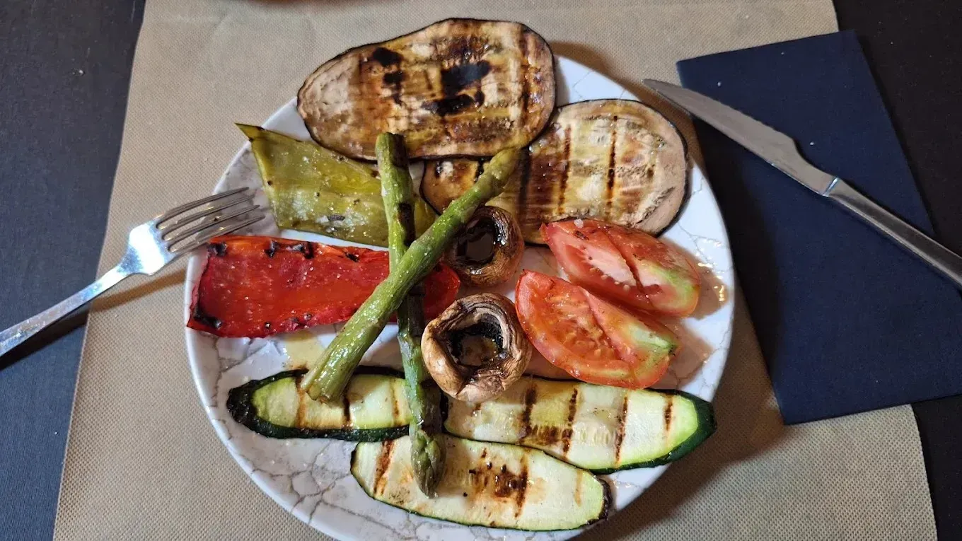 Verduras y pescado a la parrilla en un plato blanco con salsa, sobre una mesa con tenedor y servilleta.