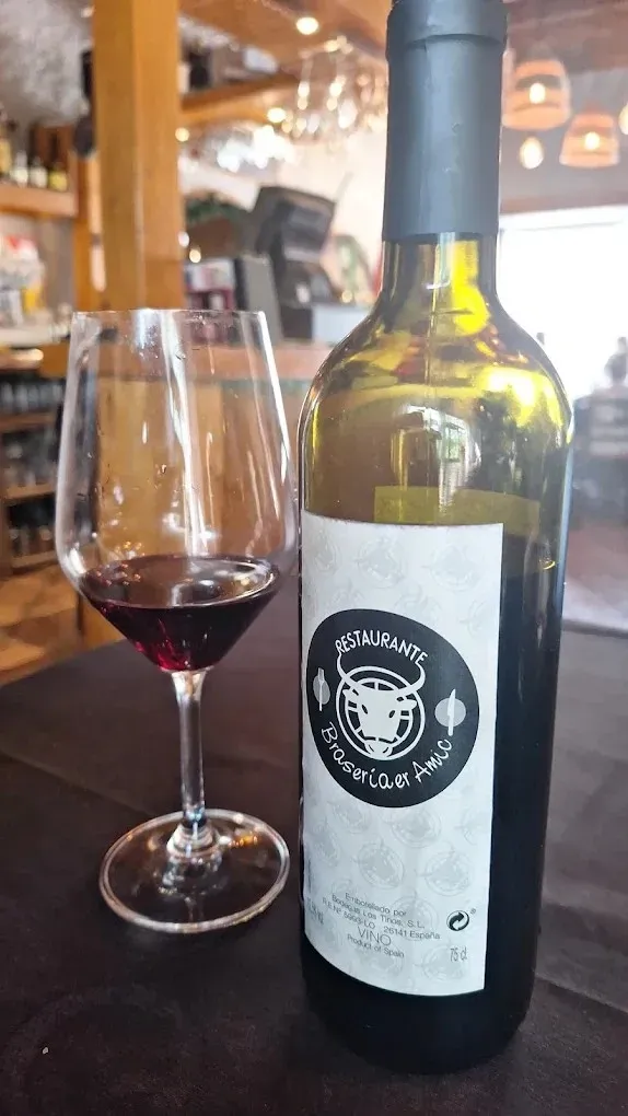 Una botella de vino junto a una copa de vino tinto sobre una mesa de madera en un restaurante con iluminación cálida.