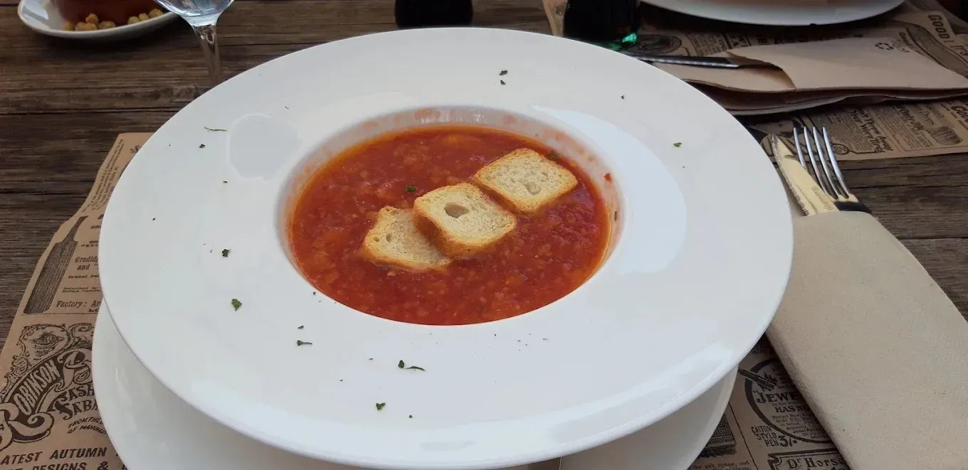 Un tazón blanco de sopa de tomate rojo con tres picatostes sobre un mantel, con vajilla al fondo.