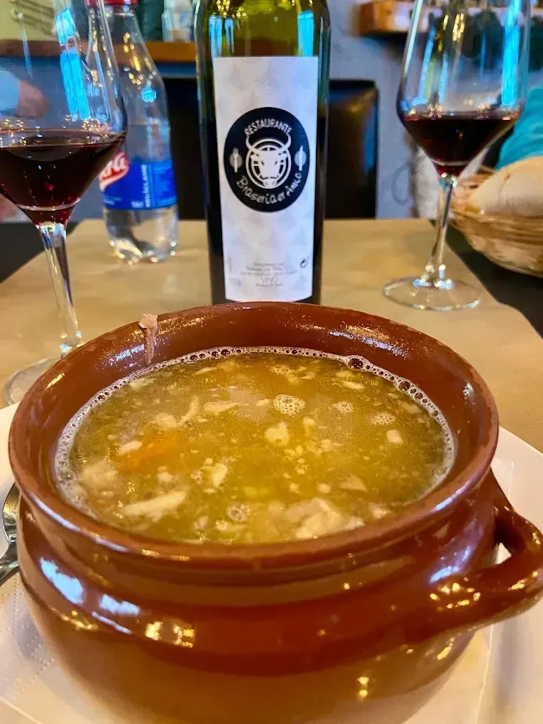 Sopa marrón en un cuenco de barro sobre la mesa de un restaurante con copas de vino y una botella al fondo.