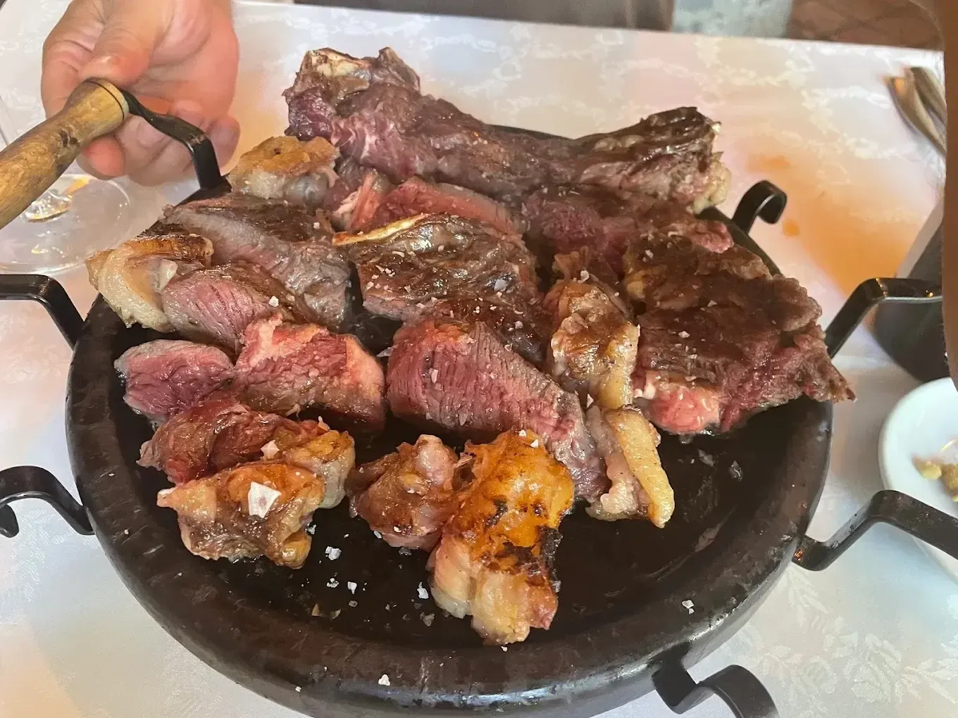 Plato de carne a la parrilla y salchichas sobre una mesa, con una mano sosteniendo un cuchillo en el borde izquierdo.