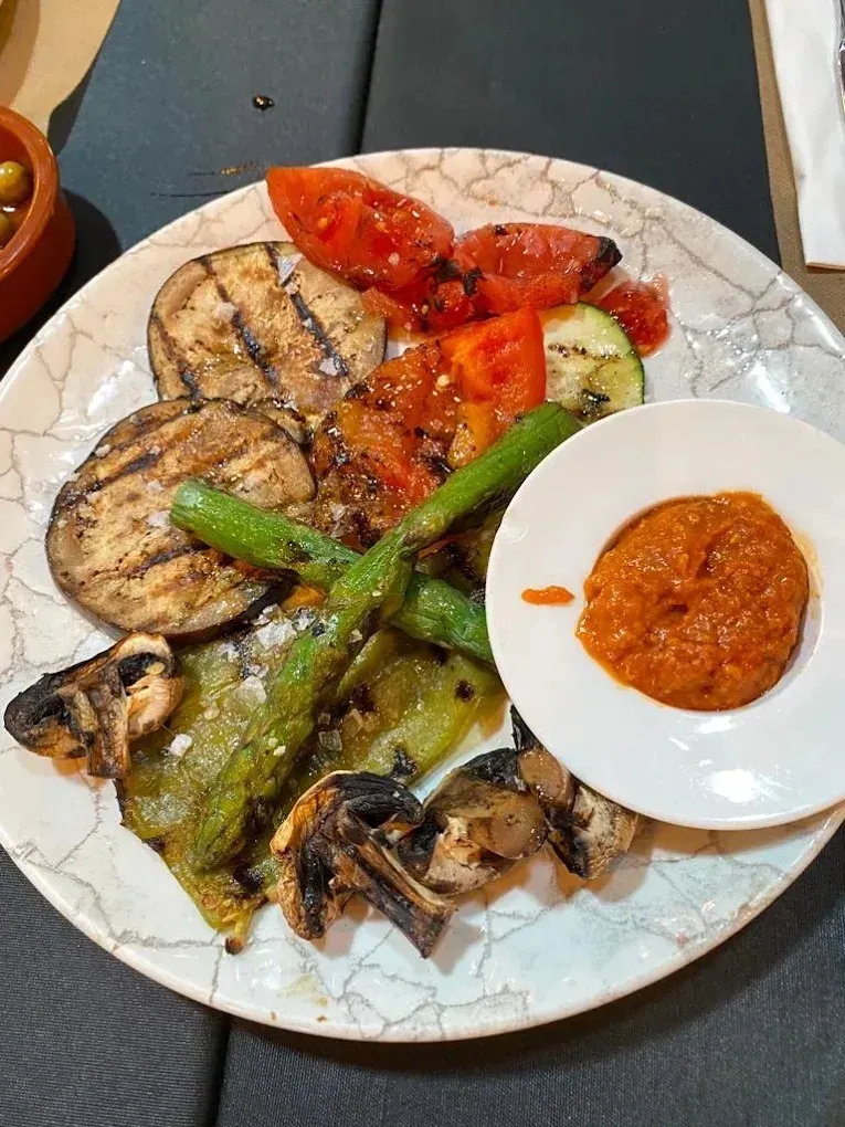 Berenjenas, calabacines, pimientos y tomates a la parrilla en un plato estampado con un pequeño cuenco de salsa roja.