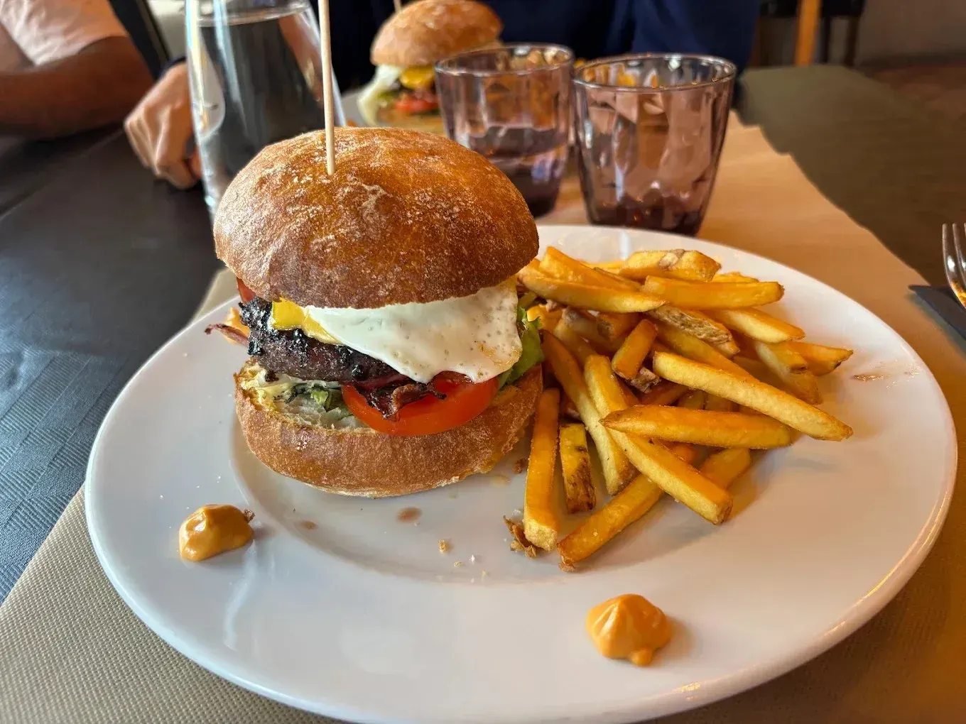 Hamburguesa y patatas fritas en un plato blanco en una mesa de restaurante con vasos al fondo.