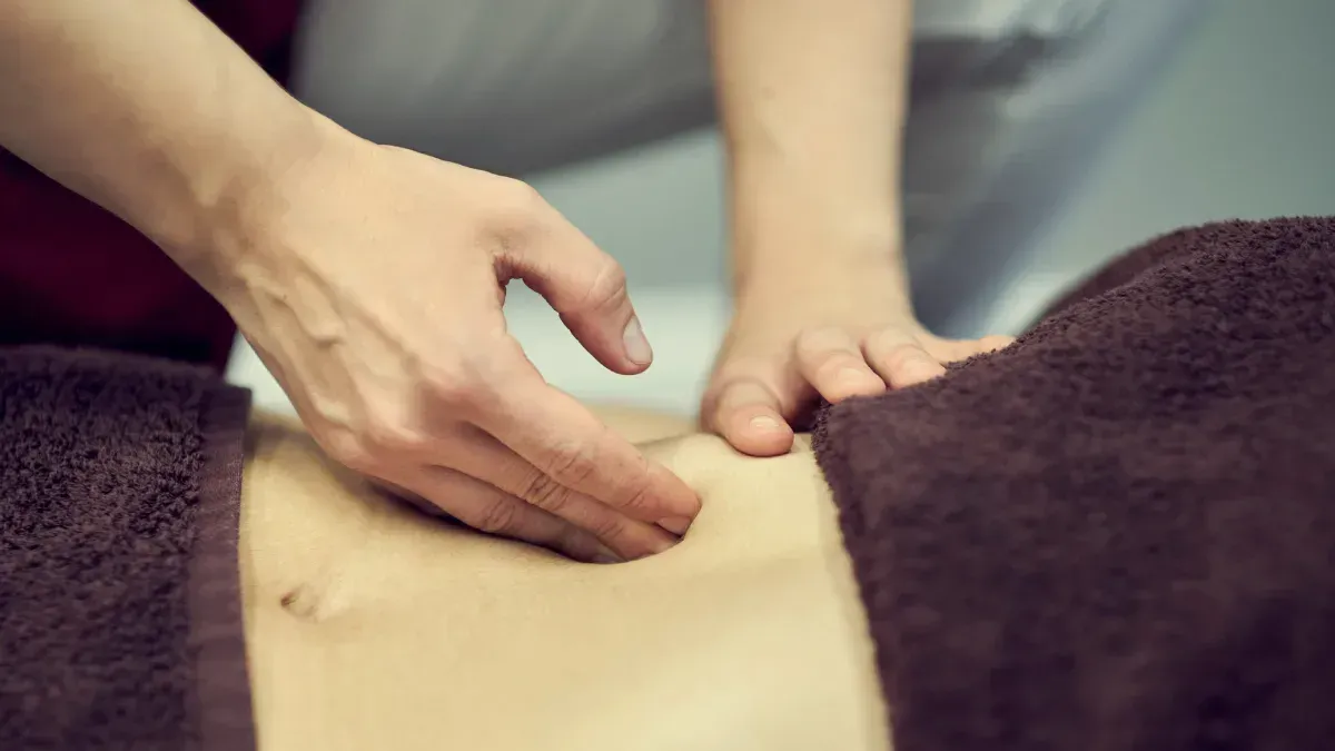 Manos dando masaje abdominal a una persona acostada, envuelta en una toalla marrón.