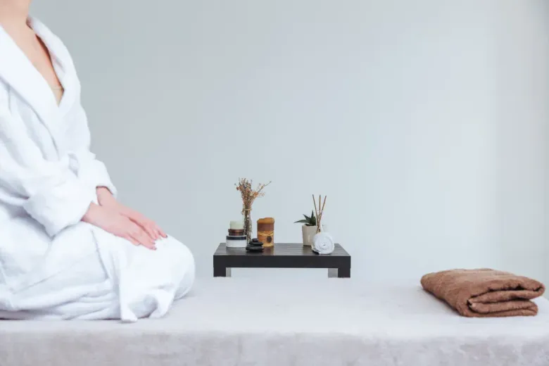Persona con una bata blanca sentada en el borde de una mesa de masajes, con artículos de spa en una bandeja