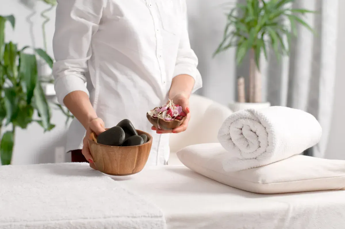 Persona preparando la mesa de tratamiento de spa con toallas, piedras y flores.