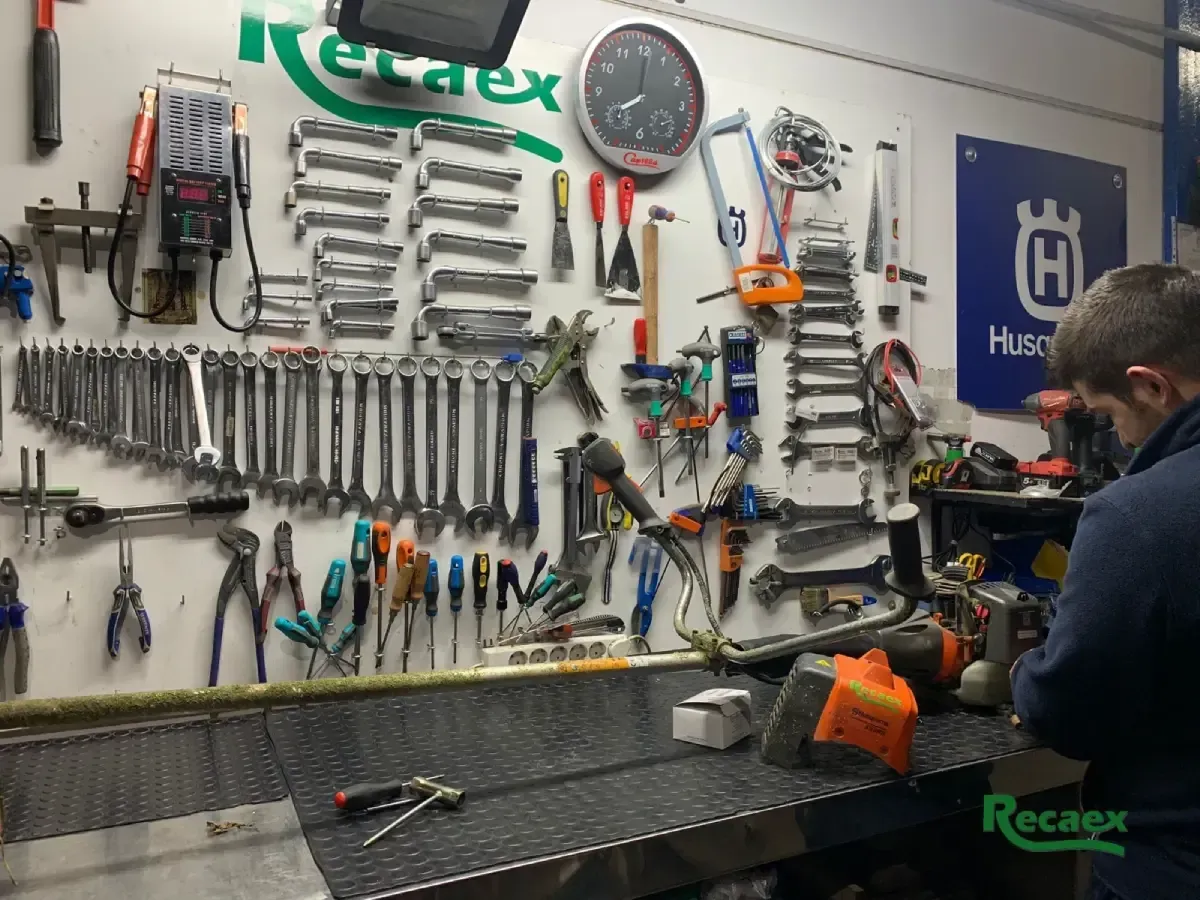 Mecánico trabajando en una motosierra naranja junto a una pared de herramientas en un taller.