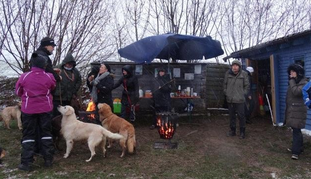 Im Freien versammelten sich Menschen um ein Feuer unter einem Sonnenschirm; in der Nähe befanden sich zwei Hunde und ein blauer Schuppen.