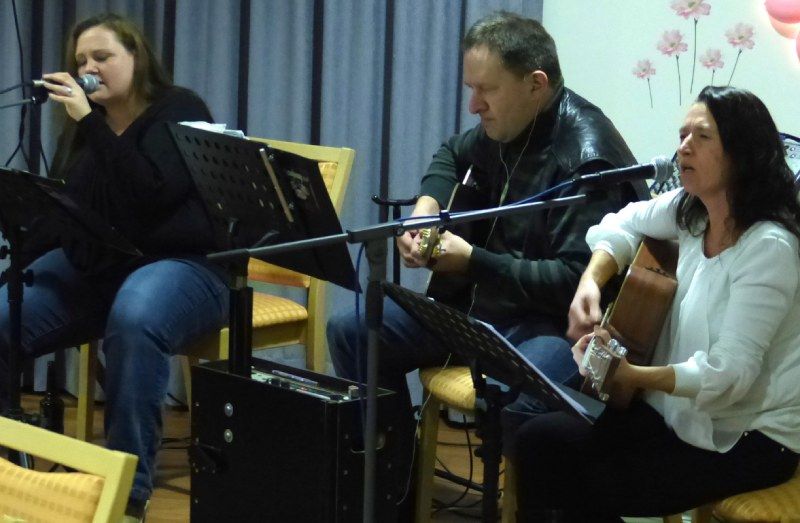 Drei Musiker treten in einem Innenraum auf, einer singt, während zwei Gitarre spielen und in Mikrofone singen.
