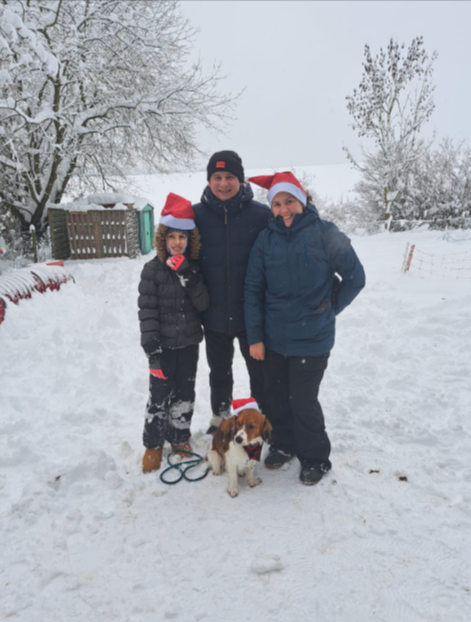 Drei Personen und ein kleiner Hund stehen auf einem verschneiten Weg, bekleidet mit Wintermänteln und Mützen.