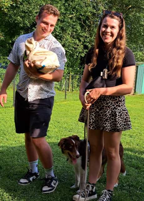 Zwei Personen stehen mit einem Hund auf einer Wiese; eine hält ein kleines Tier, die andere eine Leine.