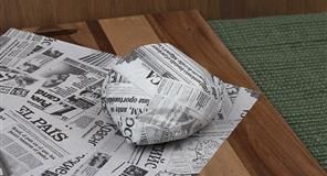 Objeto cubierto de periódico sobre una tabla de cortar de madera, al lado de una superficie texturizada de color verde.