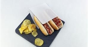 Dos perritos calientes con ketchup en una funda de papel, servidos con patatas fritas en un plato de pizarra.