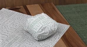 Hamburguesa envuelta en papel blanco impreso sobre una tabla de cortar de madera, al lado de un mantel verde.