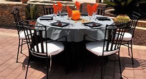 Mesa redonda dispuesta para comer con seis sillas negras, servilletas naranjas y un mantel gris en un patio.