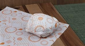 Sándwich envuelto en papel blanco con dibujos naranjas sobre una tabla de madera.