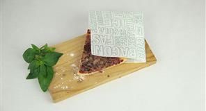 Rebanada de tocino sobre una tabla de madera con una servilleta impresa; ramita de albahaca al costado.