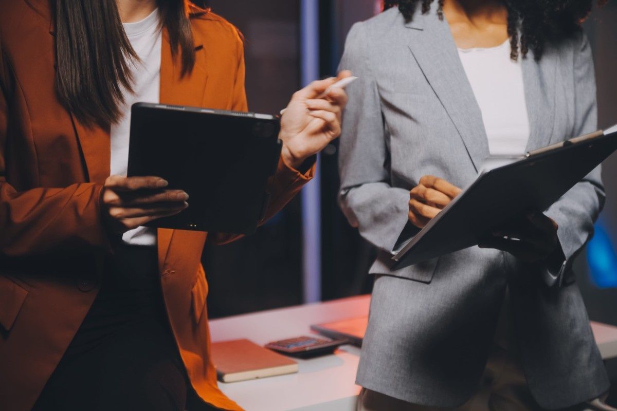Dos personas vestidas de traje de negocios están de pie en una oficina; una sostiene una tableta y hace gestos, la otra sostiene una carpeta.