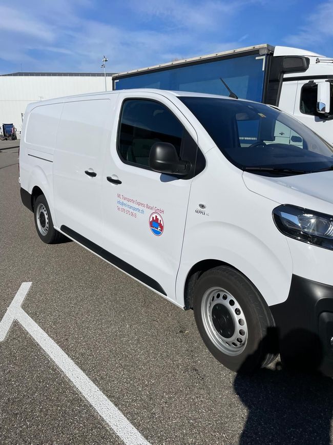 Weißer Lieferwagen mit Firmenlogo auf einem gepflasterten Parkplatz geparkt; im Hintergrund ist ein LKW-Anhänger zu sehen.