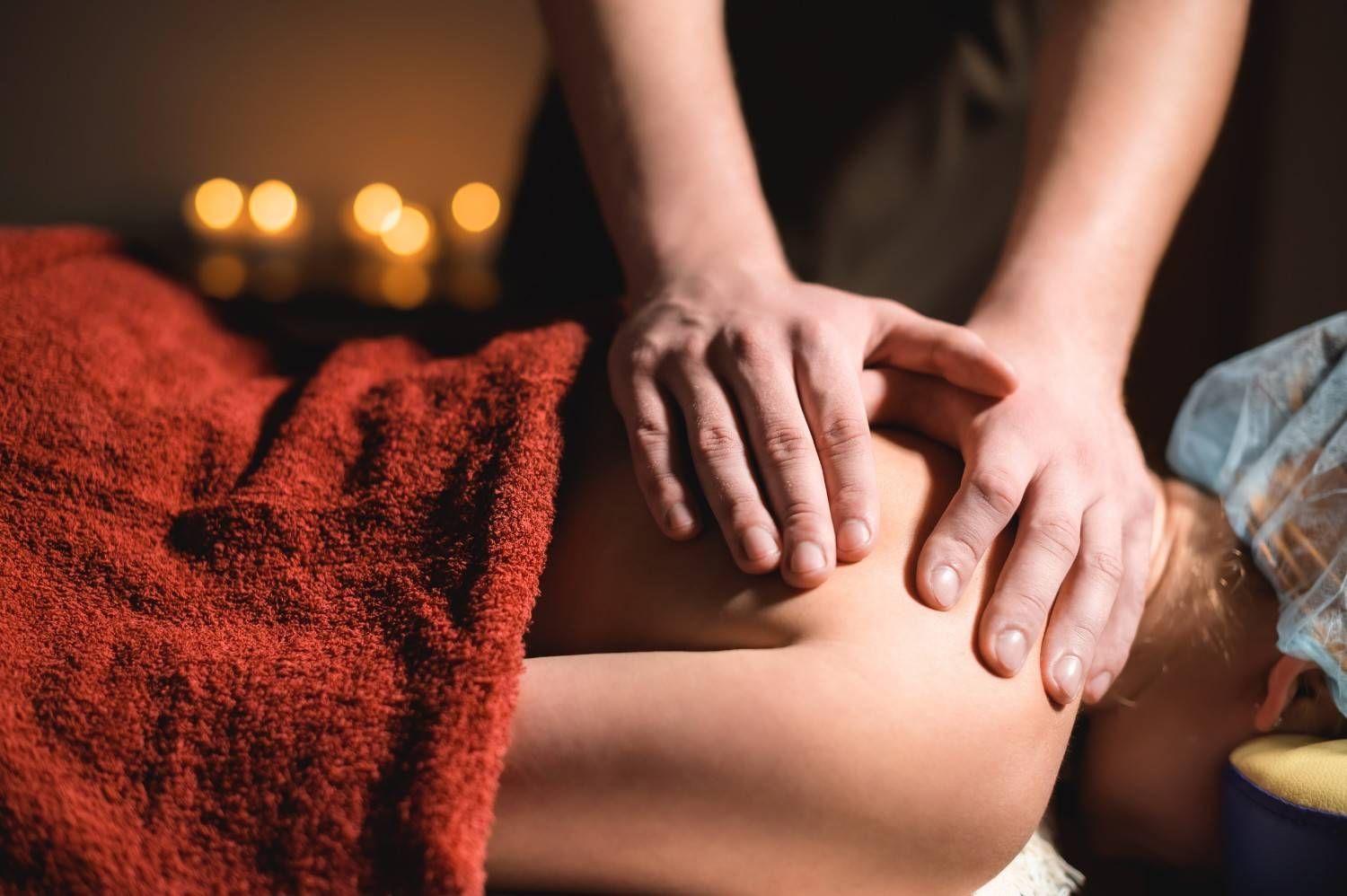 Manos masajeando el hombro de una persona, cubierto con una toalla roja, con velas en el fondo borroso.